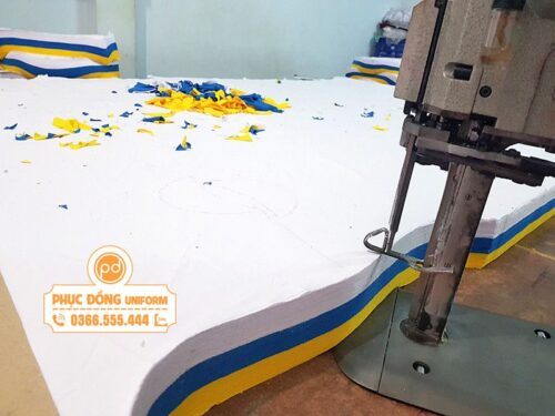 Máy cắt vải công nghiệp đang cắt bàn vải nhiều lớp trong quy trình trải vải may áo thun đồng phục